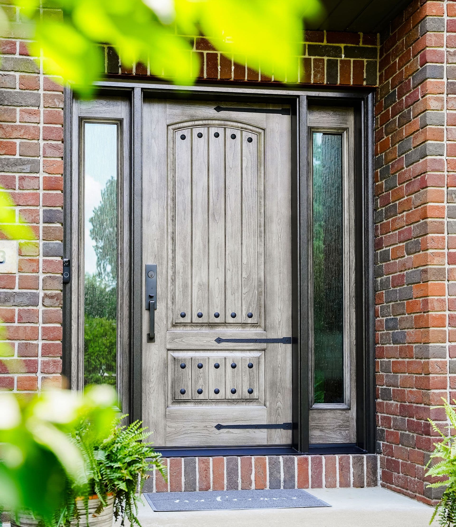 Ornate gray wooden front door with glass sidelights set in a red-brick entryway, framed by lush green leaves and ferns; welcoming, rustic tone.