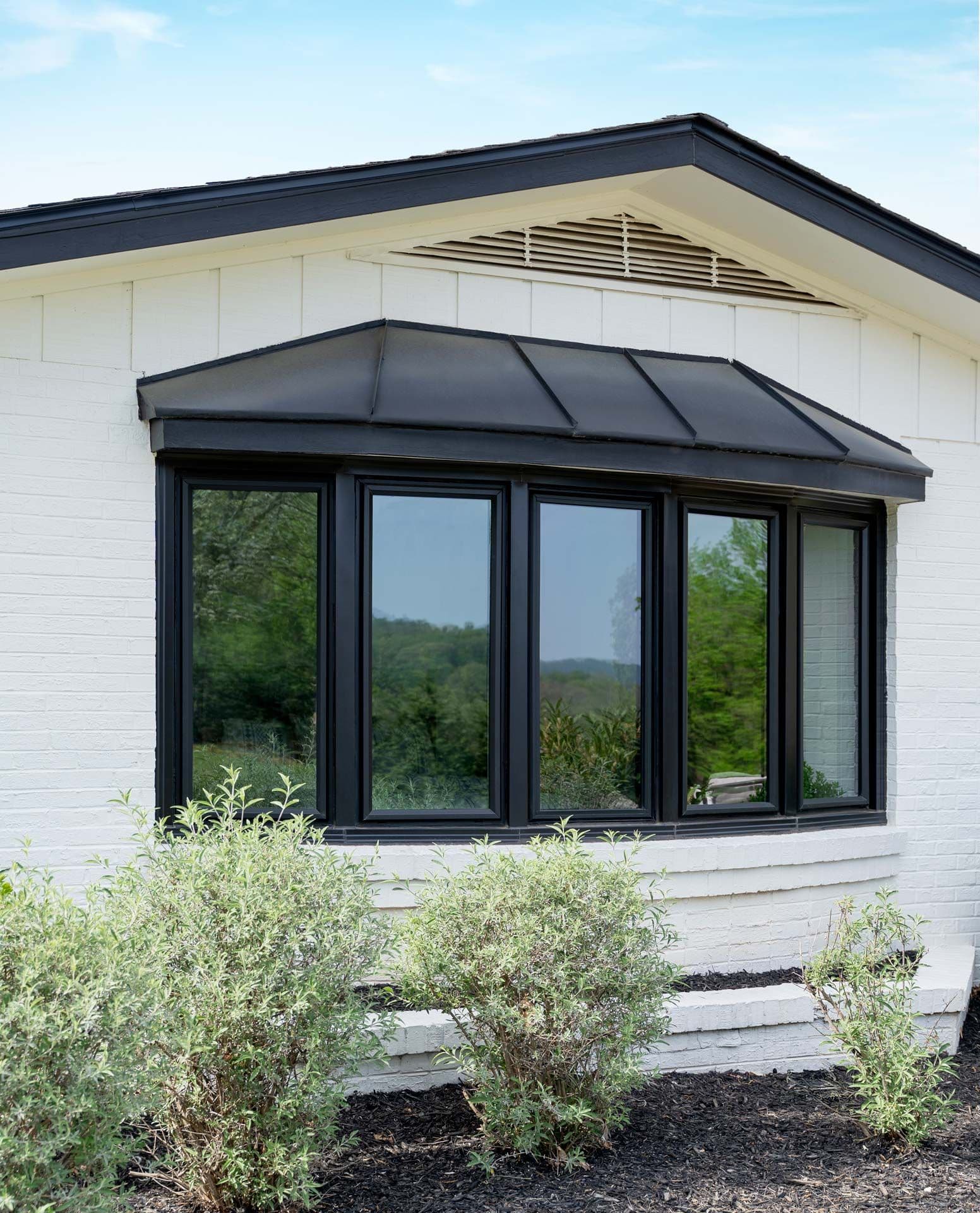 White cottage exterior with black-framed bay windows, metal awning, and neatly planted shrubs, creating a bright, inviting rural scene.