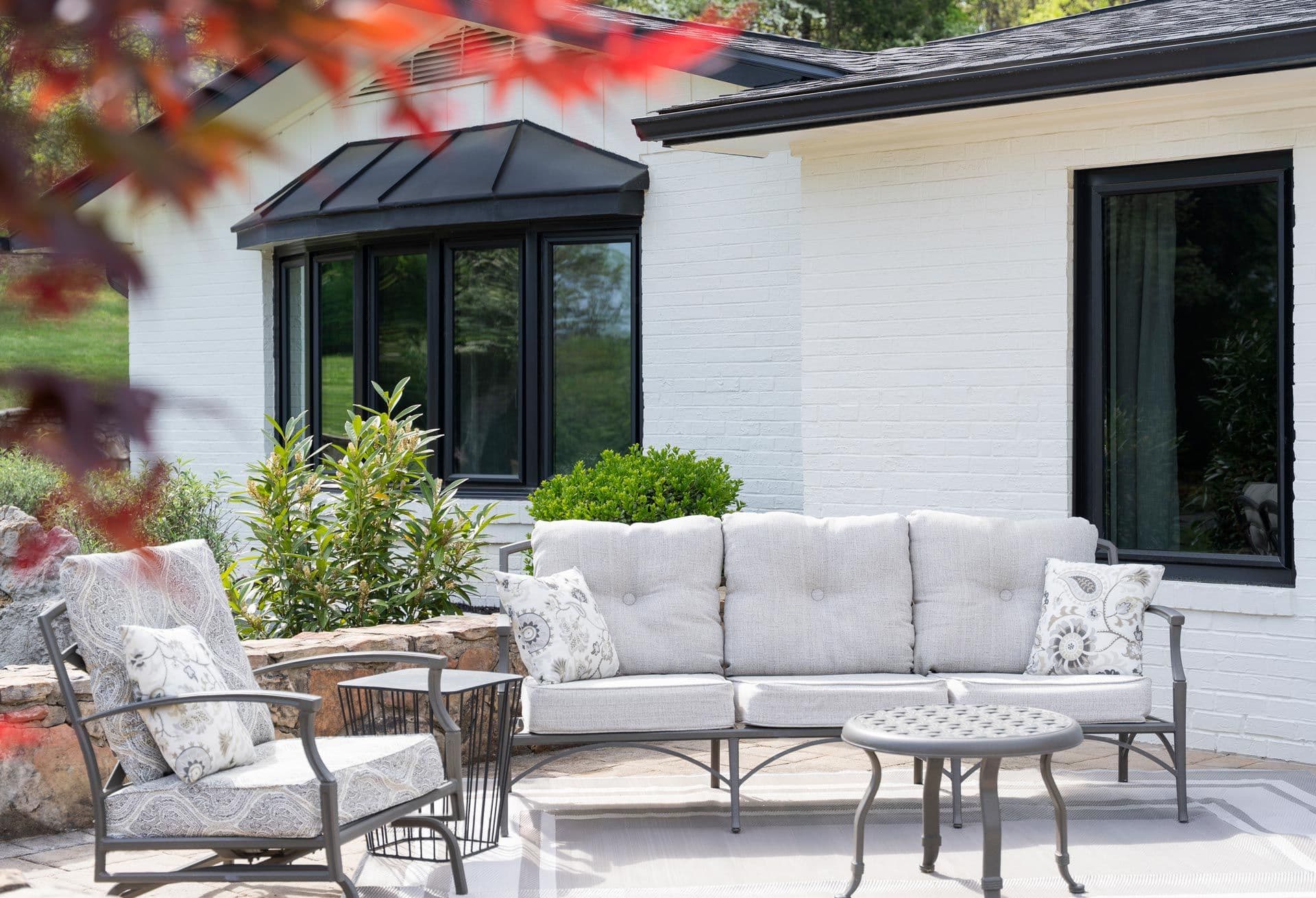 Bright, inviting patio with white brick house, cushioned sofa and chairs, plants, and glass table, framed by autumn leaves.