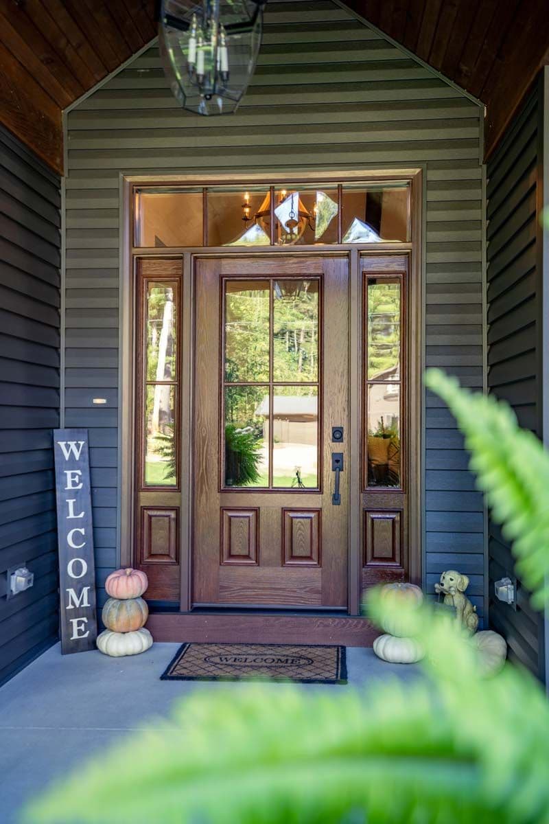 Welcoming cottage entryway with wooden double doors, pumpkins, “WELCOME” sign, hanging lantern, and lush greenery in sunlight.