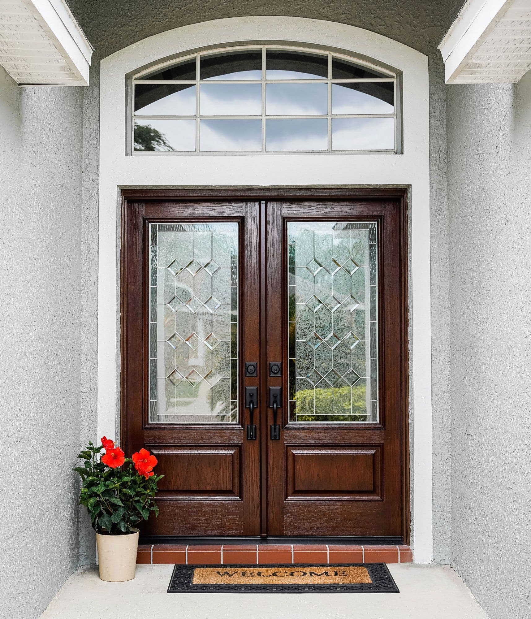 Elegant dark wood double front door with patterned glass, arched transom window, red flowers, and a welcome mat create an inviting entryway.