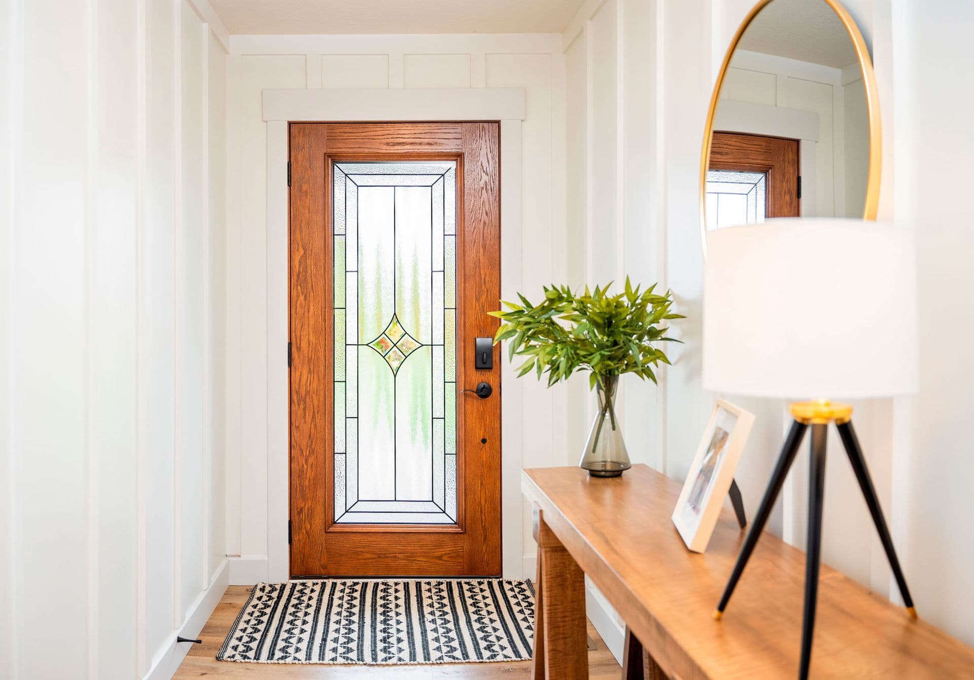 Bright, welcoming entryway with a wood stained-glass door, patterned runner, console table, leafy plant, mirror, and lamp.